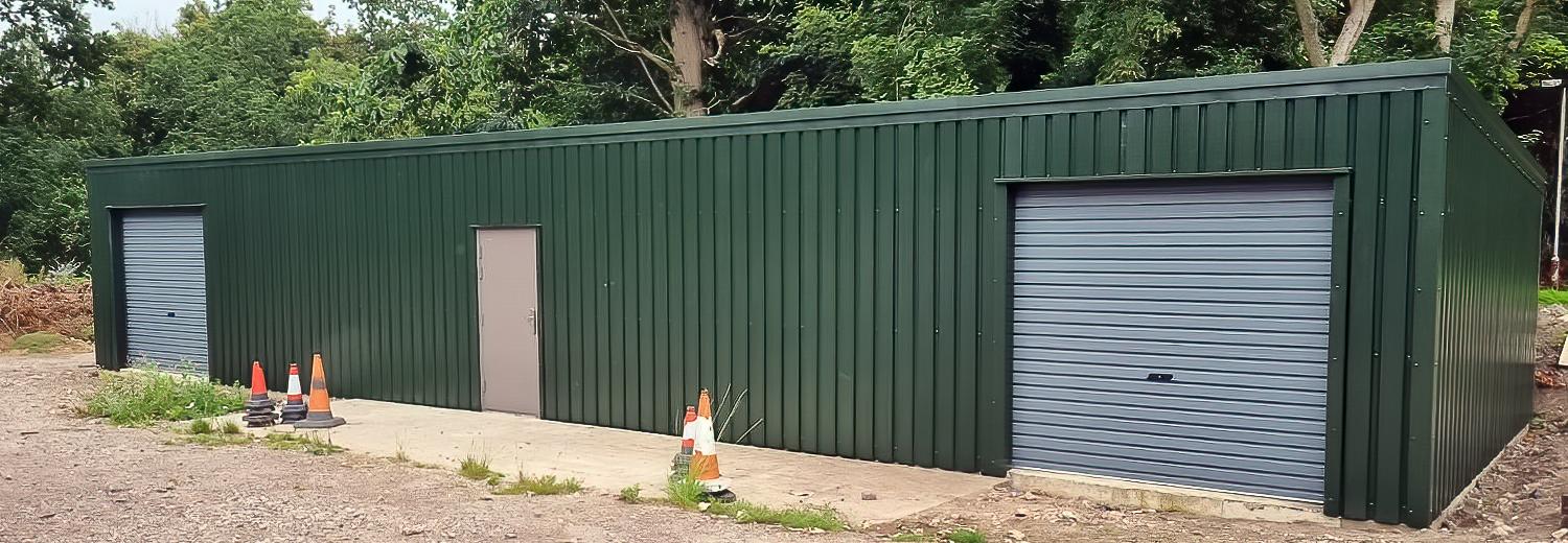 A long, panoramic view of a dark green metal utility building with two separate roller shutter doors and a central personnel door.